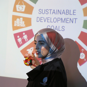 A photo of Nusaiba Talahmeh, a young women wearing a black shirt. The Sustainable Development Goals icon is showing in the background.